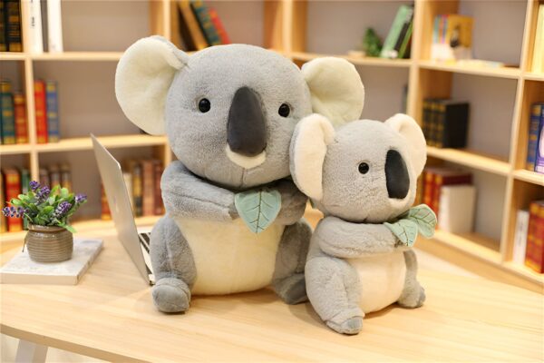 Deux adorables peluches de koala grises tenant des feuilles vertes. Les koalas sont assis sur une table en bois avec une bibliothèque en arrière-plan.