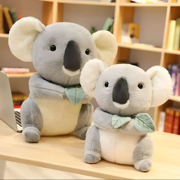 Deux adorables peluches koala, l'une tenant une feuille verte, l'autre portant un collier de feuilles vertes, posées sur une table en bois.