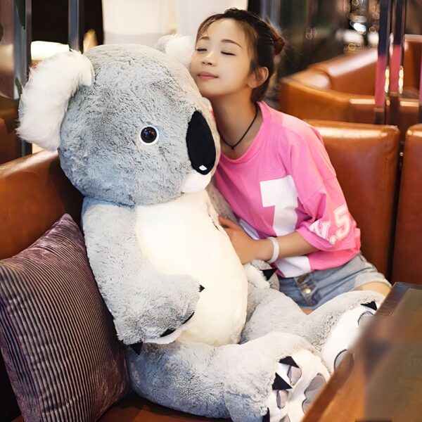 Une jeune femme portant un t-shirt rose et un short en jean est assise sur un canapé en cuir marron avec un grand ourson en peluche gris.