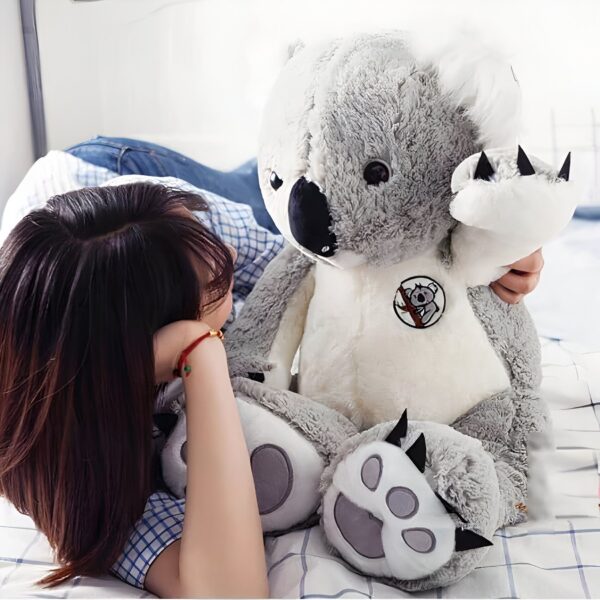 Une jeune femme allongée sur un lit avec un grand koala en peluche gris et blanc.