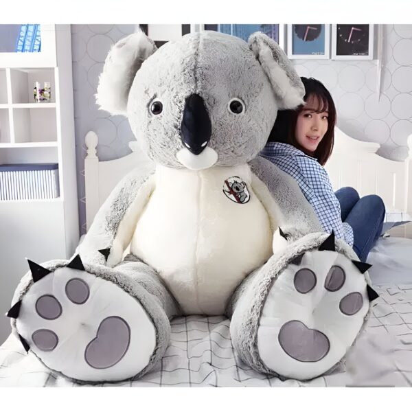 Grande peluche koala gris et blanc assis sur un lit avec une jeune femme assise derrière lui.