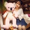 Une jeune femme souriante tenant un grand koala en peluche rose assis sur un lit. Le koala porte un badge rose et blanc avec un dessin de koala. La femme porte un chemisier blanc et une jupe en jean. Le lit est en bois et recouvert de draps blancs.