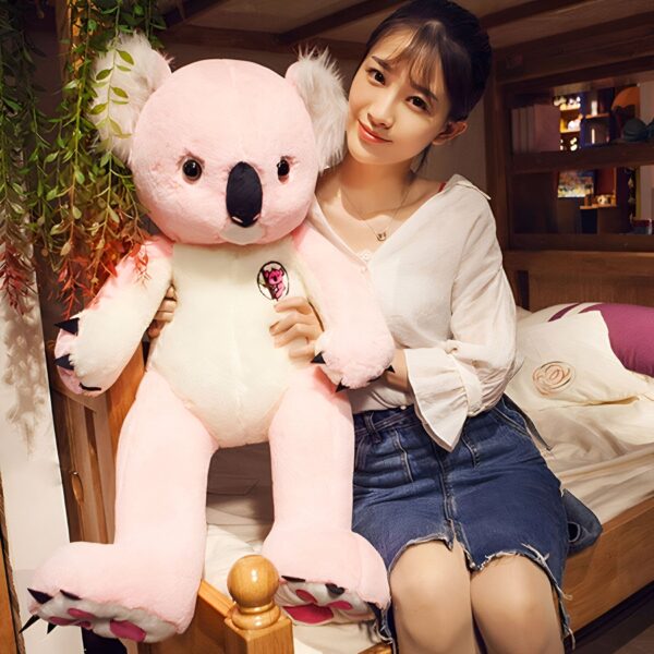 Une jeune femme souriante tenant un grand koala en peluche rose assis sur un lit. Le koala porte un badge rose et blanc avec un dessin de koala. La femme porte un chemisier blanc et une jupe en jean. Le lit est en bois et recouvert de draps blancs.