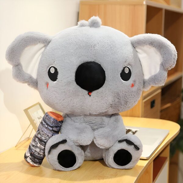 Une peluche de koala gris assise sur une table en bois avec un coussin en forme de rondin d'arbre à ses côtés.