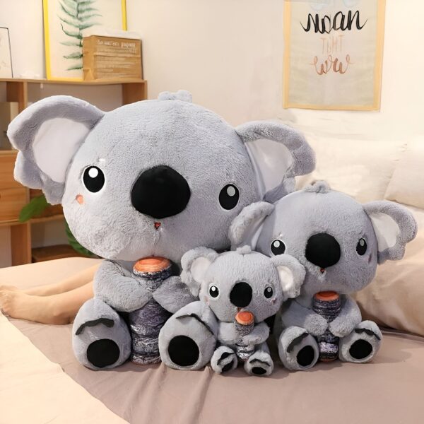 Trois peluches koalas grises assises sur un lit, tenant un morceau de bois dans leurs mains. Le plus grand koala est assis à gauche, le koala moyen est assis au milieu, et le plus petit koala est assis à droite.