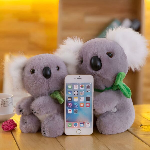Deux peluches de koala grises et douces avec un téléphone intelligent blanc entre elles.