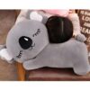 peluche-koala-grise-oreiller-doux-pour-dormir-2a Un gros ourson en peluche gris avec de grands yeux fermés et un sourire. L'ourson est allongé sur un canapé et sert d'oreiller à une personne.