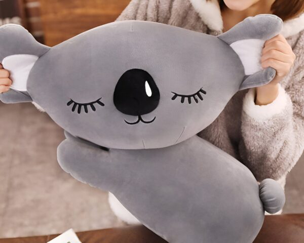 peluche-koala-grise-oreiller-doux-pour-dormir-3a Un coussin en peluche gris en forme de koala avec des yeux fermés et un nez noir.