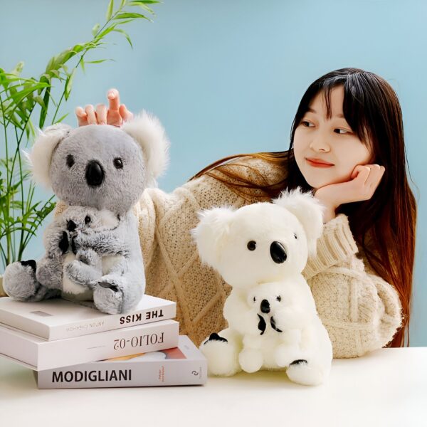 Une jeune femme souriante avec deux peluches koalas, l'une grise et l'autre blanche. Les koalas sont assis sur une table avec des livres.