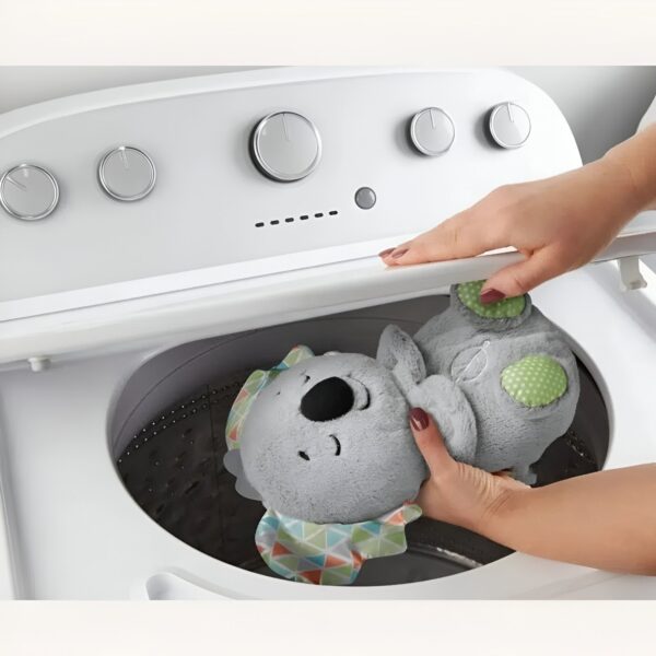 Une main place une peluche de koala dans une machine à laver. La peluche est grise avec un ventre coloré.