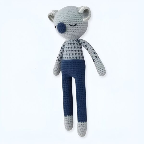 Un koala en peluche gris et bleu, crocheté, portant un pantalon bleu et un haut à motifs gris.
