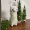 Peluche lama blanc debout devant deux petits sapins de Noël décorés de boules rouges et dorées.