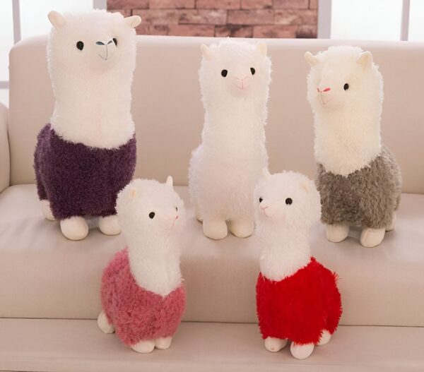 Cinq peluches de lama en position debout avec différents manteaux. Les lamas sont disposés sur un canapé blanc. Les peluches sont blanches avec des détails de couleurs différentes.