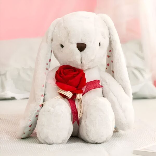 Un lapin en peluche blanc tenant une rose rouge sur un lit blanc.