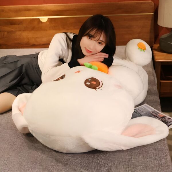 Une jeune femme souriante est allongée sur un lit avec une grande peluche en forme de lapin blanc tenant une carotte orange dans ses pattes.