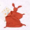 peluche-lapin-doudou-bebe-en-coton-et-mousseline-3a-3 Une couverture en tissu doux de couleur rouille avec une tête de lapin brodée. La couverture est parfaite pour les bébés et les jeunes enfants.