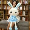 Une jeune femme tient une grande peluche de lapin blanche portant une robe bleue avec des étoiles blanches. La peluche a un nœud bleu sur la tête et le mot "Love" brodé sur une oreille.