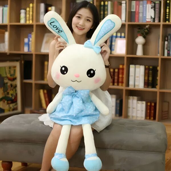 Une jeune femme tient une grande peluche de lapin blanche portant une robe bleue avec des étoiles blanches. La peluche a un nœud bleu sur la tête et le mot "Love" brodé sur une oreille.