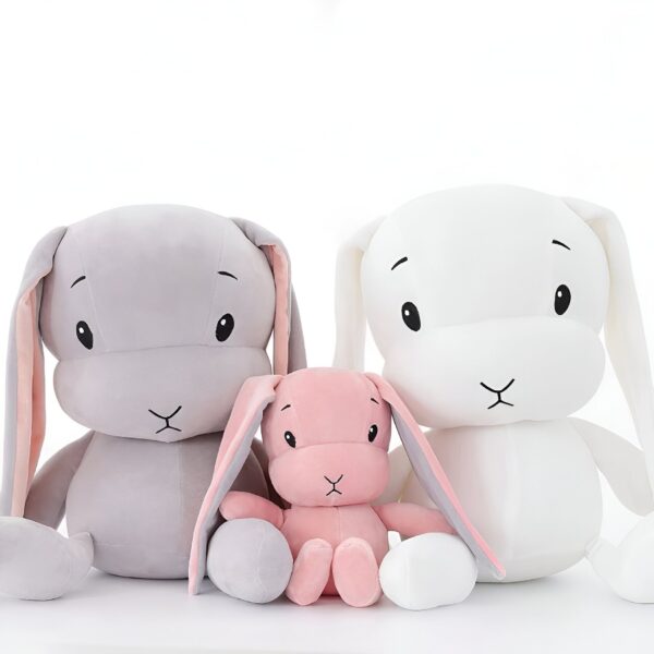 Trois lapins en peluche, un gris, un blanc et un rose, assis sur un fond blanc.