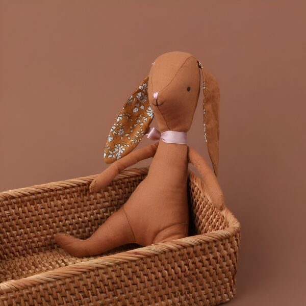 Un lapin en tissu brun avec des oreilles à fleurs jaunes et un nœud rose dans un panier en osier.