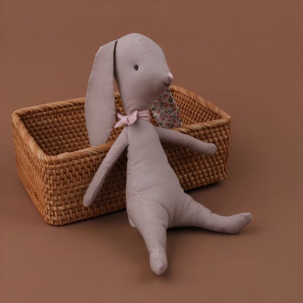 Un lapin en tissu gris avec un nœud rose est assis sur un panier en osier.