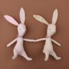 Deux lapins en peluche, un rose et un beige, se tiennent la main sur un fond brun.