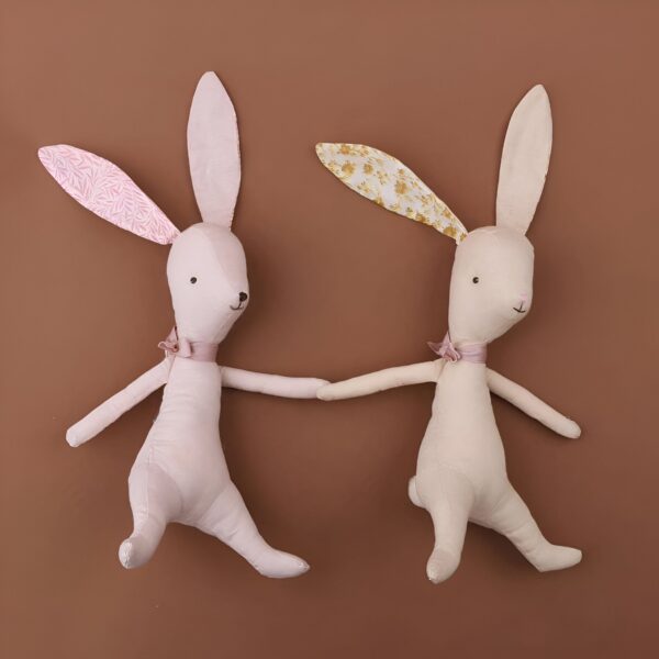 Deux lapins en peluche, un rose et un beige, se tiennent la main sur un fond brun.