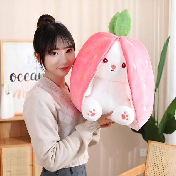 Une femme souriante tient une peluche lapin blanche en forme de fraise rose.