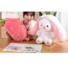Une femme tient un oreiller en forme de fraise et un lapin en peluche avec des oreilles roses.