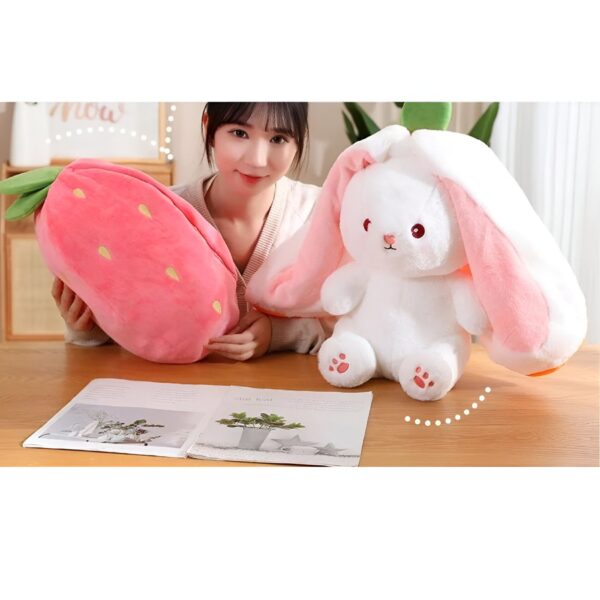 Une femme tient un oreiller en forme de fraise et un lapin en peluche avec des oreilles roses.