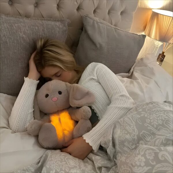 peluche-lapin-respirant-doudou-lumineux-apaisant-pour-sommeil-profond-2a-2 Une jeune femme dort dans son lit avec un lapin en peluche lumineux. Le lapin a des grandes oreilles et une lumière douce et chaude. Elle est confortablement installée dans sa literie avec des oreillers et des couvertures moelleuses.