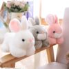 peluche-lapin-rose-tout-doux-2a Trois lapins en peluche, un blanc, un gris et un rose, sont assis sur un banc en bois.