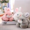 peluche-lapin-rose-tout-doux-3a Trois peluches en forme de lapin, un rose, un blanc et un gris, posées sur un canapé blanc.