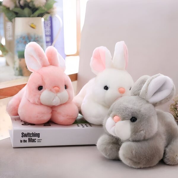 peluche-lapin-rose-tout-doux-3a Trois peluches en forme de lapin, un rose, un blanc et un gris, posées sur un canapé blanc.