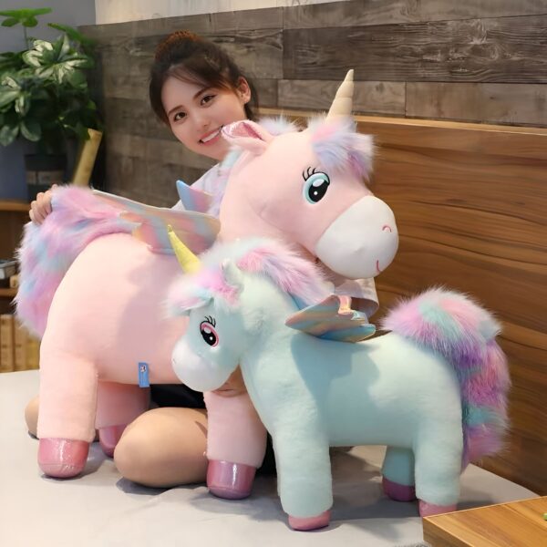 Une jeune femme souriante tient deux peluches licornes, une rose avec des ailes arc-en-ciel et une bleue avec une crinière arc-en-ciel.