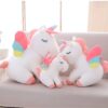 Trois peluches en forme de licorne blanche avec des ailes arc-en-ciel, assises sur un canapé rose.