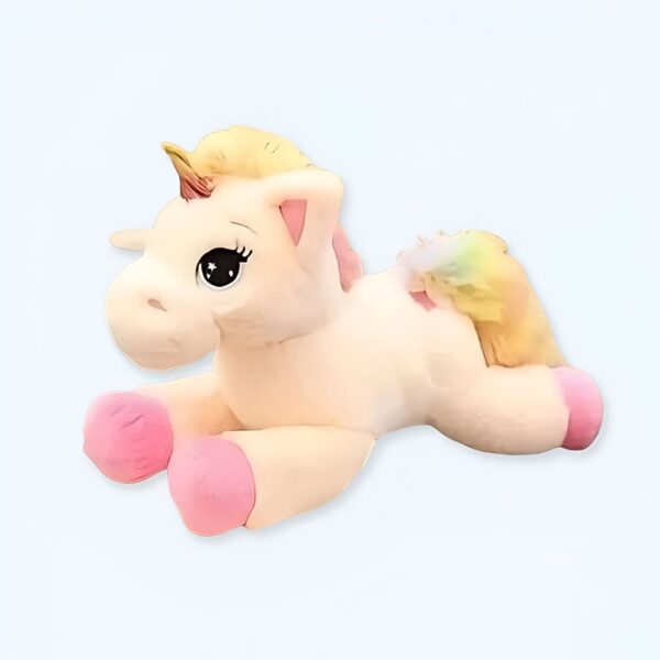 Une peluche de licorne blanche avec une crinière jaune, une queue arc-en-ciel et des yeux noirs. Elle est couchée sur le ventre.