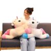 Une jeune femme souriante est assise sur un canapé avec deux grandes peluches de licorne. Les licornes sont blanches avec des crinières arc-en-ciel et des sabots roses et bleus.