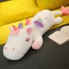 Une peluche de licorne blanche couchée sur un canapé avec une aile arc-en-ciel et le mot "Sweet" écrit sur son dos.