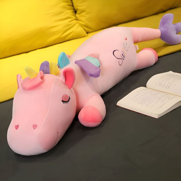 Une licorne rose en peluche avec des ailes arc-en-ciel allongée sur un canapé jaune. L'inscription "Sweet" est brodée sur son corps.