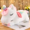 Une peluche de licorne blanche avec une corne arc-en-ciel, des yeux fermés et des ailes roses.