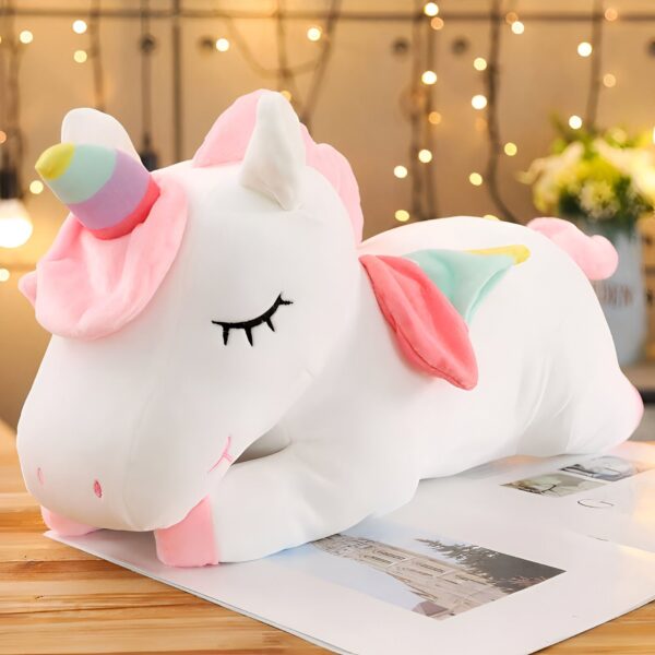 Une peluche de licorne blanche avec une corne arc-en-ciel, des yeux fermés et des ailes roses.