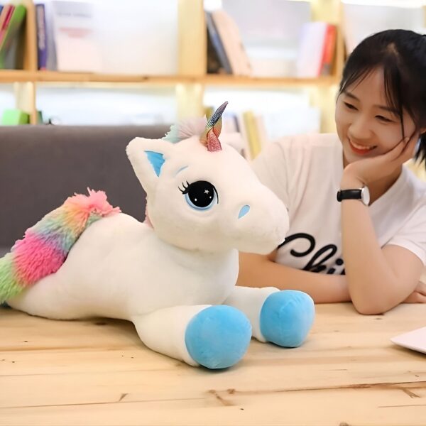 peluche-licorne-bleue-arc-en-ciel-grande-taille-cadeau-enfant-3a Une peluche de licorne blanche avec une crinière arc-en-ciel et des sabots bleus est posée sur une table en bois. Une jeune fille souriante est assise à côté de la peluche.