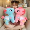 Une jeune femme assise sur un canapé, tenant deux peluches en forme de licorne, l'une bleue et l'autre rose, avec des ailes et des étoiles.