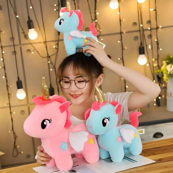 Une jeune femme souriante tient trois peluches en forme de licorne. La licorne du milieu est rose avec des ailes blanches et des étoiles jaunes. La licorne de gauche est bleue avec des ailes blanches et des étoiles jaunes. La licorne du haut est bleue et tient un chapeau avec une étoile.