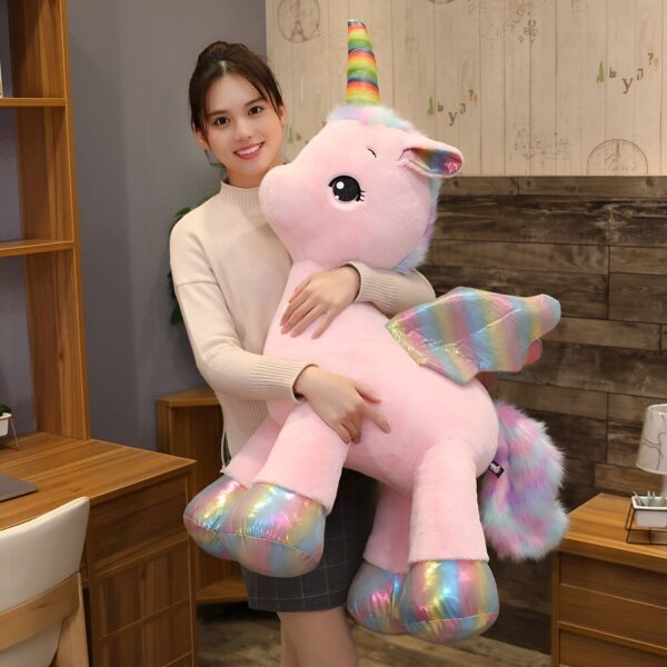 Une jeune femme tient une grande peluche rose en forme de licorne avec une corne arc-en-ciel. La licorne a des ailes arc-en-ciel et une queue de fourrure arc-en-ciel.