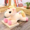 Une peluche de licorne blanche avec une crinière et une queue arc-en-ciel, allongée sur un livre sur une table en bois.