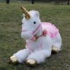 Une grande licorne en peluche blanche avec une corne dorée, un licol rose et une selle rose avec des détails de fleurs. Elle est posée sur de l'herbe verte.