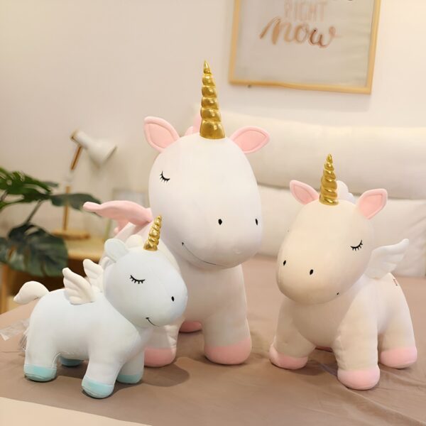 Trois peluches de licorne, une grande blanche, une moyenne rose et une petite bleue, avec des ailes et des cornes dorées.