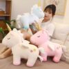 Trois peluches licornes, une bleue, une beige et une rose, avec des ailes et des étoiles. Une jeune femme sourit en tenant la licorne bleue.